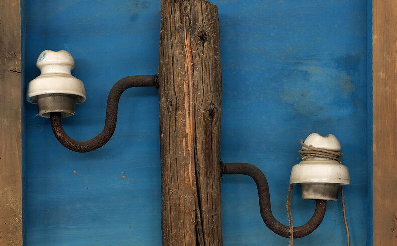 Materialbild, zentral vor einem gemalten blauen Himmel das obere Stück eines alten Strommastes mit drei Porzellanisolatoren und herunterhängenden Leitungen